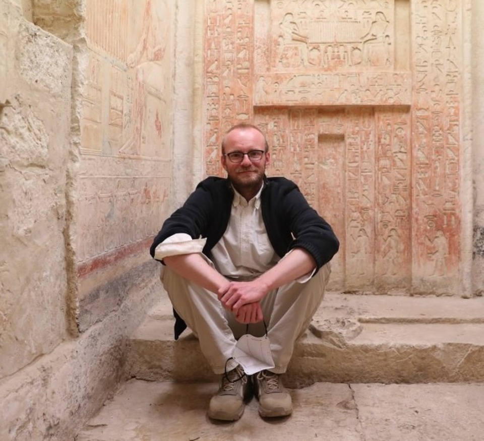 A person sitting in a squatting position with arms crossed over the knees, in front of an ancient wall covered with hieroglyphics.